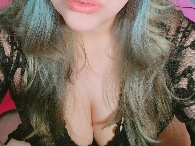 Akkanee's tits are showing through a black lace top, with her pink lips and long, wavy, blue-green hair in the foreground. Her cleavage is prominent and sexy.