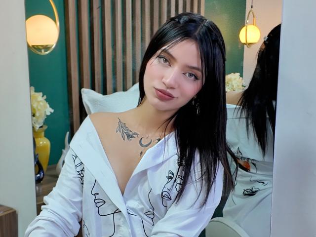 ScarlettPreatty with black hair, white off-shoulder shirt, and chest tattoo, showing tits and a subtle smile, standing in front of a mirror, green wall in background.