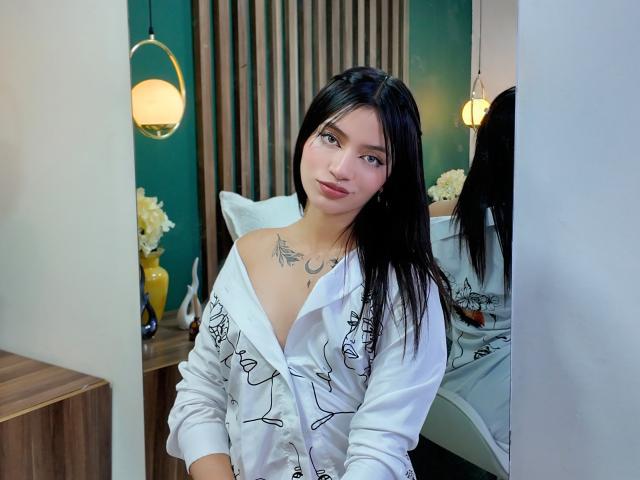 ScarlettPreatty with black hair, white off-shoulder shirt, and chest tattoo, looking at camera, showing small tits, green eyes, and subtle makeup. Mirror reflection visible.