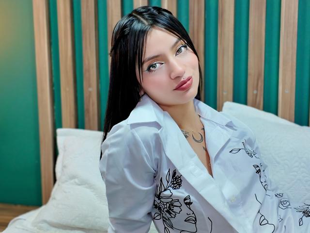 ScarlettPreatty with long black hair, blue eyes, and pink lips, wearing a white floral shirt, leaning on white pillows, her small tits and chest tattoo visible.