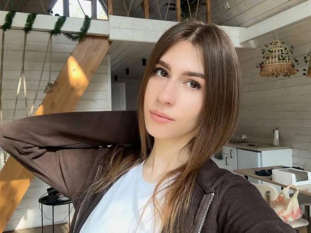 BridgettCatignani with brown hair, light skin, and brown eyes, wearing a brown zip-up hoodie over a white shirt, hand behind head, subtle smile, natural light, wooden structure.