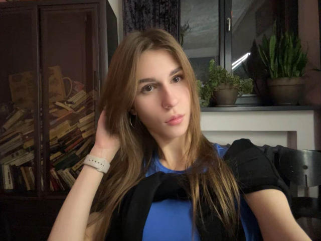 BridgettCatignani with long brown hair, blue shirt, black sleeves, white wristwatch, hand in hair, pouty lips, brown eyes, sitting, bookshelf, plants, window, daytime, indoor.