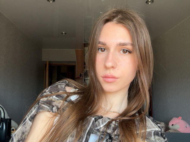 BridgettCatignani has long brown hair, brown eyes, and a neutral expression. She's wearing a camouflage shirt, and a small gold necklace is visible. Her ass is not shown.