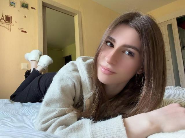 BridgettCatignani, lying on a bed, face down, with her ass up, wearing a white hoodie and black pants, brown eyes, long brown hair, small hoop earrings, smiling.