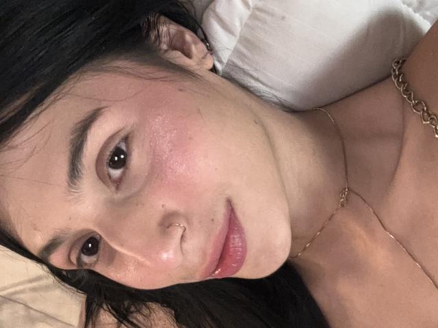 SophieBeaumont has a cute face with pinkish cheeks, brown eyes, and a nose ring. She's lying down with black hair, wearing a gold necklace. Boobs partially visible.