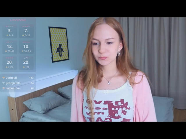NenitaTraino mit langen, roten Haaren, die einen rosafarbenen Pullover über einem weißen Tanktop mit roter Schrift trägt, steht in einem Schlafzimmer und blickt mit einem leichten Lächeln nach unten..