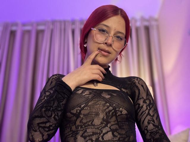 CherryMist with red hair, wearing glasses, black lace top, finger on lips, looking down, purple lighting, pale skin, slight smile, cute, sexy.