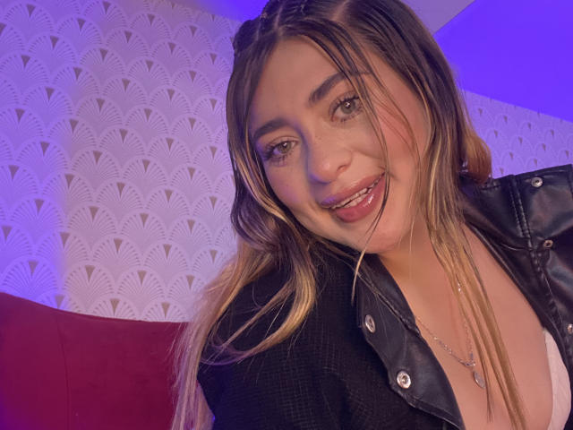 Sarah Feith mit blonden Haaren, einer schwarzen Lederjacke und einem weißen Hemd, lächelnd mit herausgesteckter Zunge, wobei ihre Zähne zu sehen sind, und Sommersprossen im Gesicht, wirkt sie süß und attraktiv..