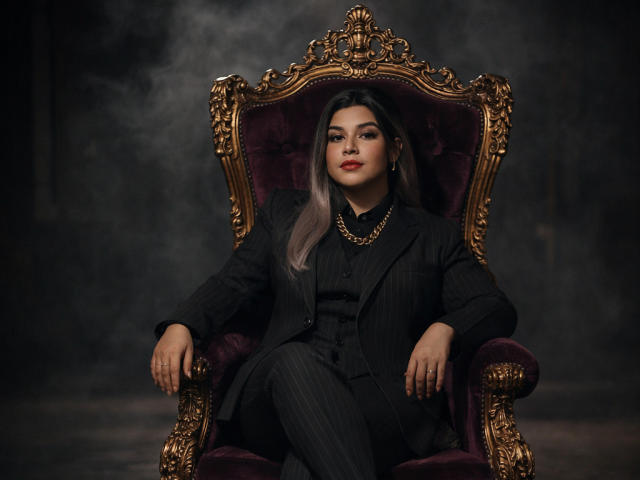 CamilaFetishDirty with big ass, sitting on an ornate gold and purple chair, wearing a black pinstripe suit, gold necklace, and red lipstick, in a smoky dark room.