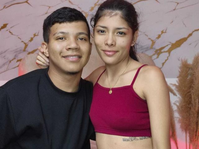 MiaHero with short black hair, black shirt, smiling, arm around MiaHero with long brown hair, red sports bra, small tits, necklace, and tattoo, standing closely.