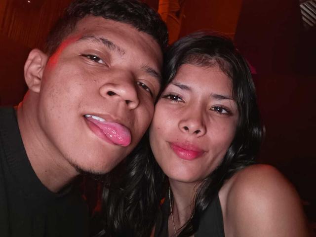 MiaHero with short brown hair and a goatee, tongue out, next to MiaHero with black hair and pink lipstick, both smiling. Red light in the background.