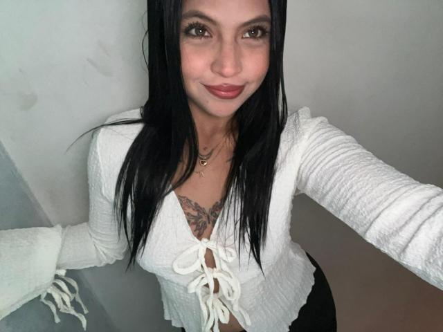 NyraVanel has black hair, brown eyes, and a small smile. She's wearing a white, lace-up, long-sleeve top that shows her tits. She's standing with one arm raised.