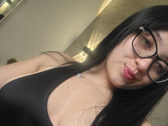 LunaBellaHotty with long black hair, glasses, and pink lipstick, wearing a black tank top showing her boobs, has a small necklace and a slight smile.