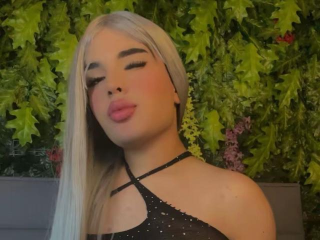 GlossyyBratz has platinum blonde hair, pink lipstick, and a black halter top with a crisscross strap. Her eyes are closed, pouting with slightly flushed cheeks.