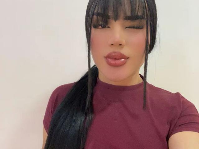 GlossyyBratz with black bangs, winking, wearing a maroon t-shirt, has glossy pink lips, and a long black ponytail, showing a cute and sexy face.