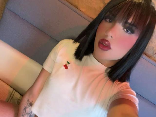 GlossyyBratz with dark bob, pink lipstick, and white t-shirt with cherry embroidery, sitting with legs apart, showing a small tattoo on her thigh.