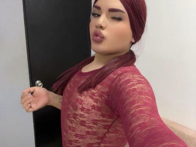 GlossyyBratz with maroon lace top, pouty lips, and long maroon hair, showing off her tits through semi-transparent fabric, standing near a black door.