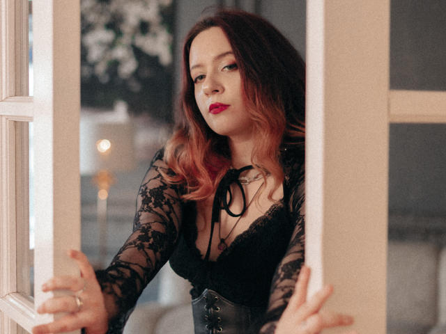 NanaMoonlight with wavy pink and brown hair, wearing black lace lingerie and corset, standing in a doorway, holding frames, red lipstick, confident expression, soft lighting.