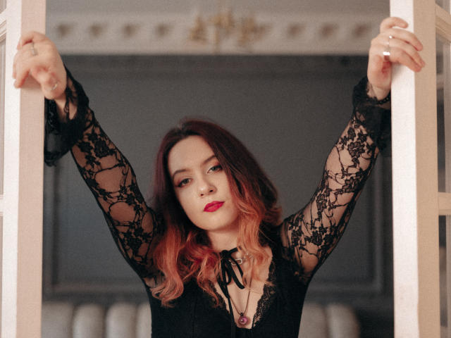 NanaMoonlight with arms raised, wearing a black lace long-sleeve top, red lipstick, and pink-to-brown ombre hair, standing between white door frames, looking directly at the camera.