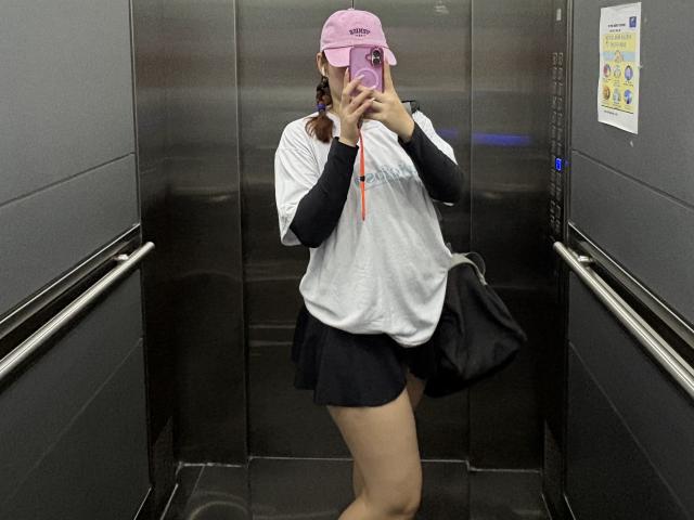 MalenaJones in a elevator, wearing a pink cap, white shirt, black skirt, and black bag, holding a phone, covering her face, with her ass and tits visible.