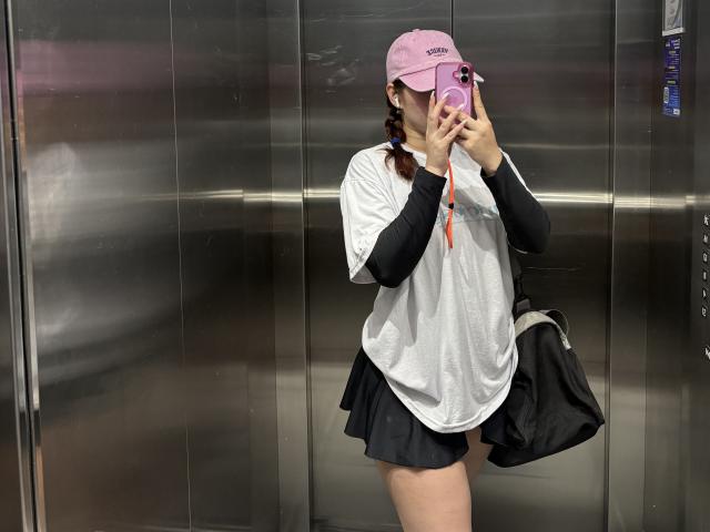 MalenaJones in a mirror selfie wearing a pink cap, white mesh shirt, black long sleeves, black shorts, holding a phone covering her face, with a black bag on her side.