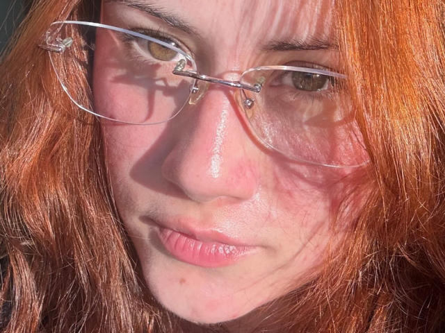 MalenaJones with red hair, clear glasses, and slightly puffy pink lips, showing a subtle smirk. Her brown eyes are focused, and sunlight casts shadows on her face.