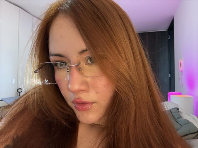 MalenaJones has long red hair, clear glasses, and pink glossy lips. She's looking slightly to the side with a serious expression. Her nose is small and cute.