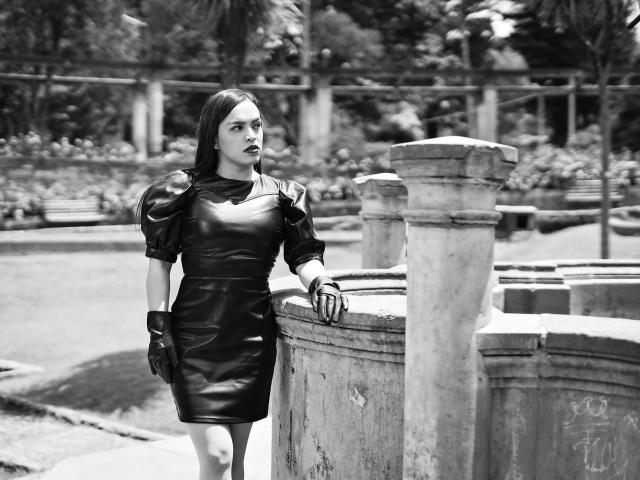 KattiyEvans está de pie, vestida con un vestido de cuero negro con mangas abullonadas, usando guantes, apoyada en una pared de piedra, con una expresión seria, lápiz labial negro, cabello corto castaño, en blanco y negro, sexy..