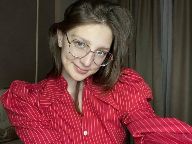 StaceyAverbach with glasses, red pinstripe shirt, smiling, medium tits, brown hair, fair skin, slight messy look, indoors, soft lighting.