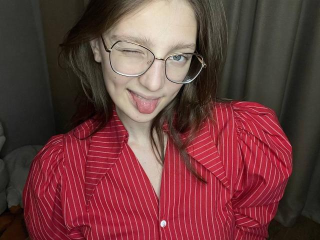 StaceyAverbach with glasses, winking, and sticking out her tongue, wearing a red pinstriped shirt, has pale skin and brown hair. Her tits are small, visible under the shirt.