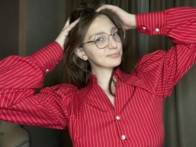 StaceyAverbach with glasses, red pinstriped shirt, and hands in brown hair, smiling slightly, showing small tits, cute, and attractive.