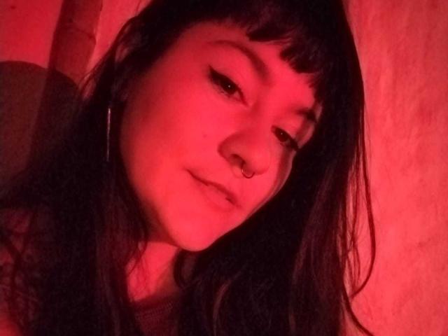 NaniSky has red-tinged skin, long black hair, and a nose ring. She's looking down with a slight smile, wearing a red top, and has a ring on her right ear.