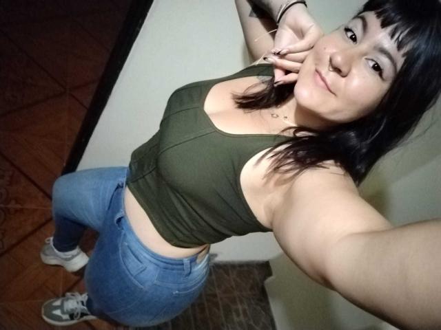 NaniSky with medium tits, green tank top, blue jeans, and white sneakers, holding up a peace sign, smiling, dark hair, standing in a room with wooden floor.