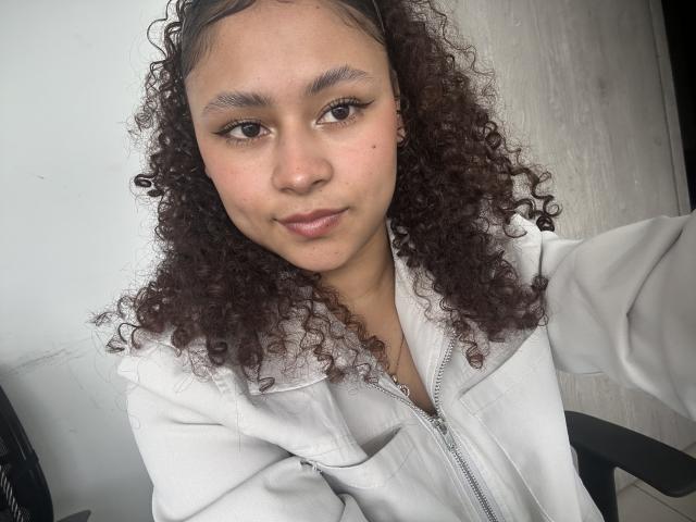 SofiaPassionne with curly brown hair, light brown skin, and a white zip-up jacket, looking at the camera with a slight smile, brown eyes, and subtle makeup.