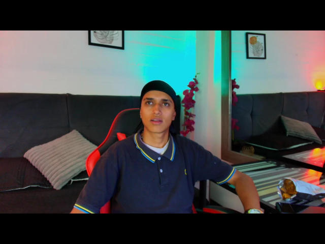 AlanColthon, wearing a black cap and navy polo with green trim, sits in a red and black gaming chair, looking slightly confused, with a mirror behind him.