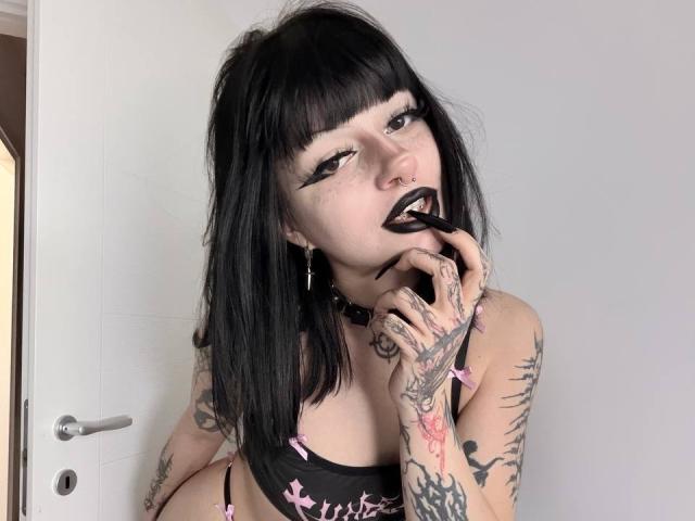 DashaBlack, avec ses cheveux noirs, son maquillage gothique, son rouge à lèvres noir, ses tatouages, son soutien-gorge rose, un doigt dans la bouche, devant une porte blanche, regardant la caméra, montrant ses seins, avec de nombreux tatouages sur les mains et les bras..