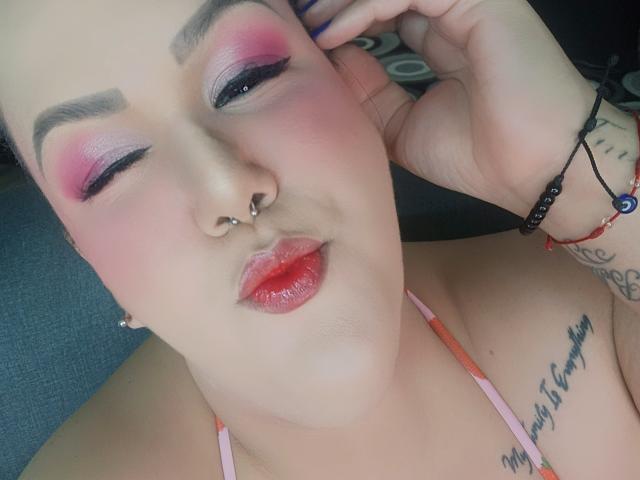 GiiannaMorrys has pink eyeshadow, red lipstick, nose piercing, and a septum ring, with a tattoo under her right breast, winking with one eye, touching her head.