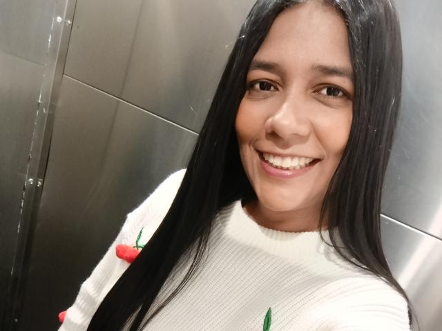 PauletteLoveX with long black hair, smiling, wearing a white sweater with red cherry designs, in a metallic elevator, has light brown skin and dark brown eyes.