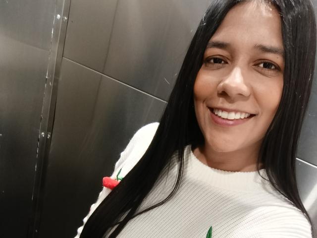 PauletteLoveX with long black hair, smiling, wearing a white sweater with green and red details, in a metallic elevator, showing her cute face and small tits.