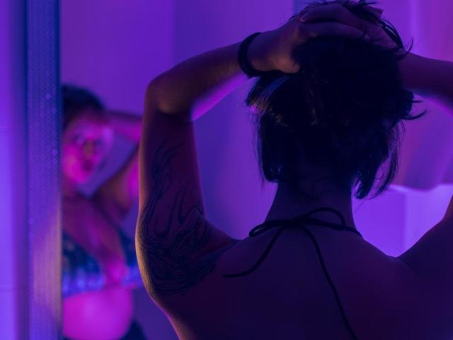 CatBleu with arms up, back to camera, purple lighting, tattoo on arm, wearing black string bikini, mirror reflection shows smiling face, pink nose, black hair, purple light.