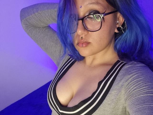 DelyBlack with blue hair, glasses, nose ring, and lip ring, wearing a grey top with black and white striped trim, showing her tits. Purple light in background.