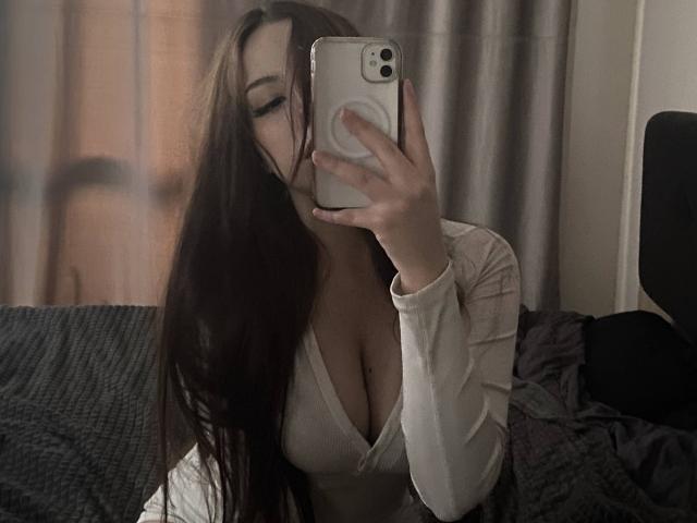TaniaRaffa with long brown hair, taking a mirror selfie, showing her cleavage in a white long-sleeve top, holding an iPhone, with a soft, dimly lit bedroom background.