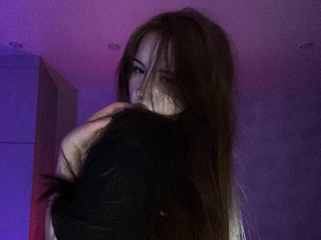 TaniaRaffa with long brown hair, partially covering her face, holding a black-haired person's head against her chest, under purple and blue lighting.