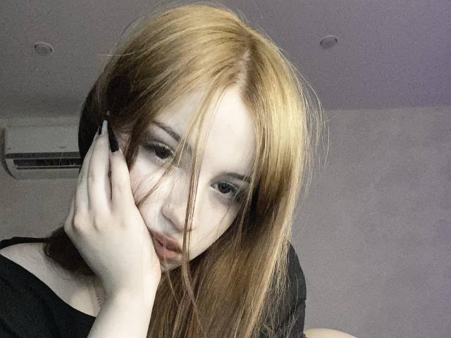 TaniaRaffa with long, messy red-brown hair, pale skin, and dark eyes, hand on face, looking down, wearing black shirt, subtle makeup, air conditioner in background.