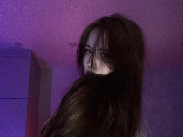 TaniaRaffa's face partially obscured by long brown hair, looking over her shoulder with a slightly mysterious, almost seductive expression, in dim purple-lit room.