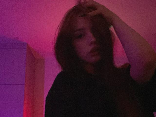 TaniaRaffa with dark, messy hair, raising her right hand, has a moody expression, wearing a black top, under pink and purple lighting. Her tits are barely visible.