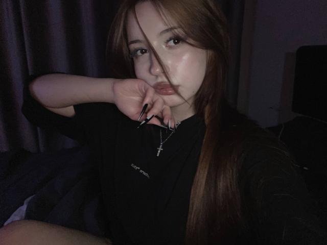 TaniaRaffa with long brown hair, black nails, black shirt, cross necklace, biting finger, dark room, dim light, serious expression, pale skin, small ass, slim figure.