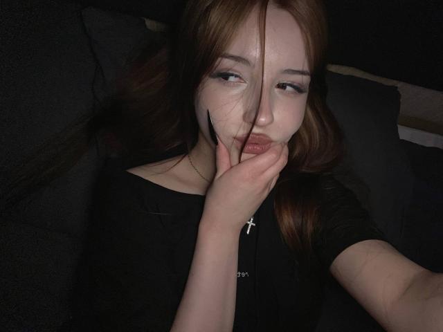 TaniaRaffa with long brown hair, black shirt, and cross necklace, biting her hand, pouty lips, half-lidded eyes, pale skin, dark background, hand covering lower face.