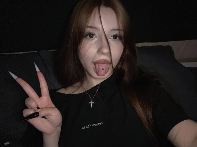 TaniaRaffa with long brown hair, black shirt, tongue pierced, sticking tongue out, making a peace sign with long nails, wearing a cross necklace, dark background.