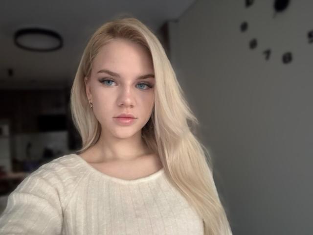 AviciAura has blonde hair, blue eyes, and wears a white ribbed sweater. Her tits are small and her ass is not visible. She looks straight at the camera.